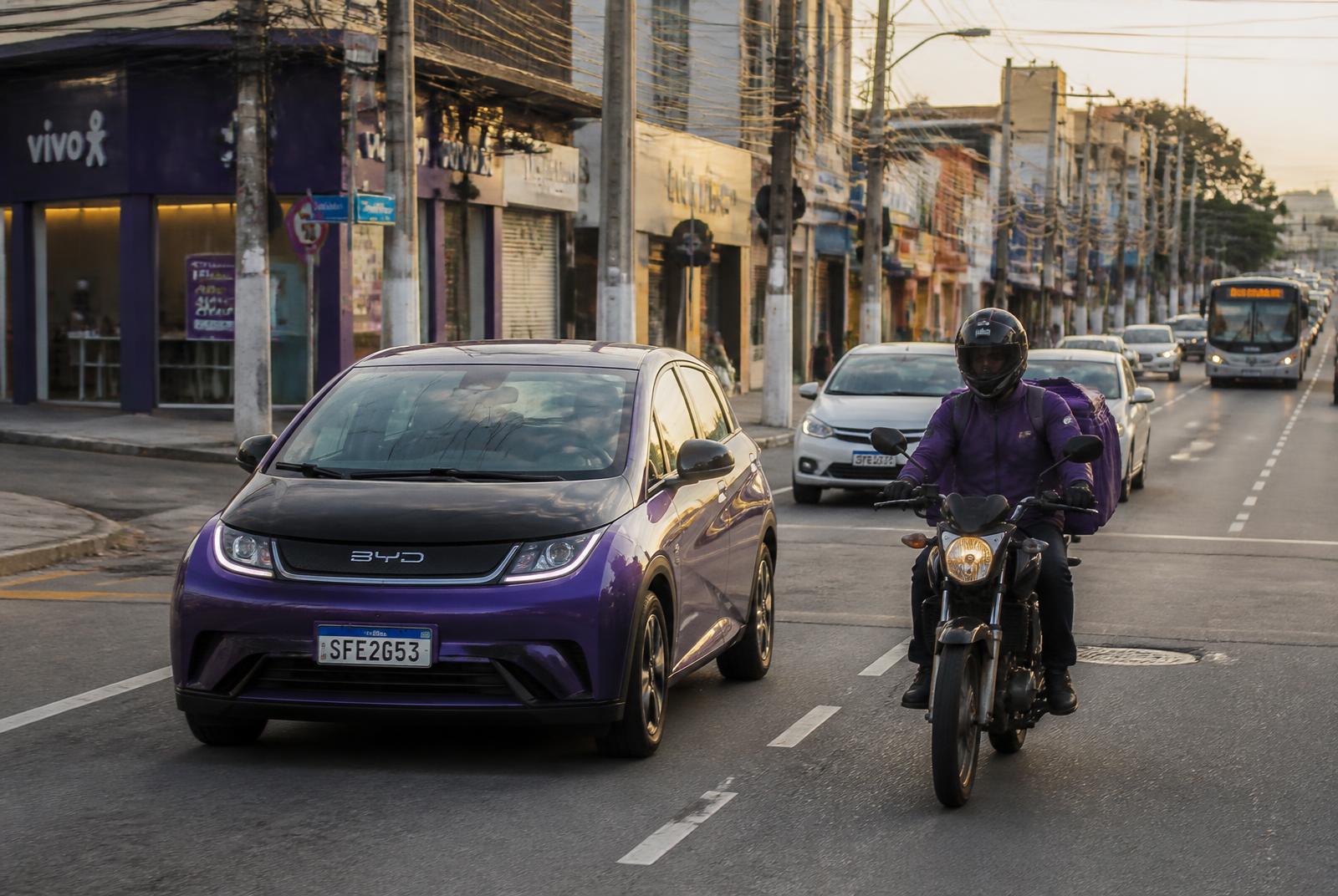 Carro elétrico roxo e entregador de moto circulando por uma avenida da cidade