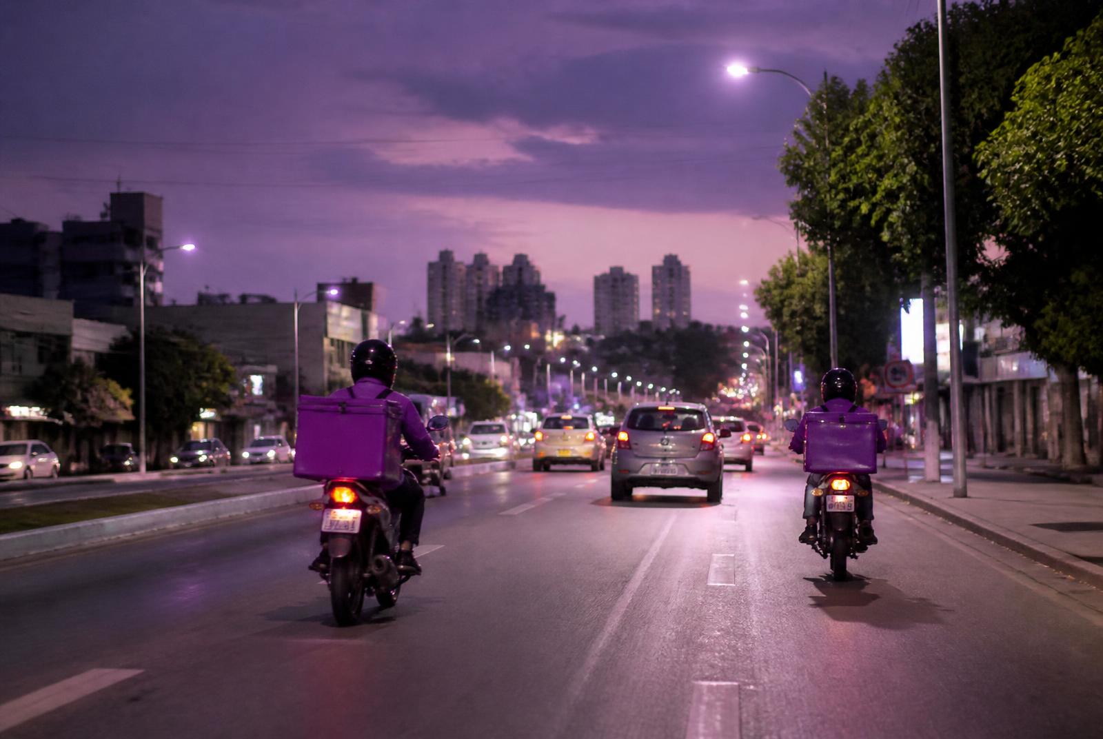 Avenida movimentada ao entardecer com carros e entregadores de moto
